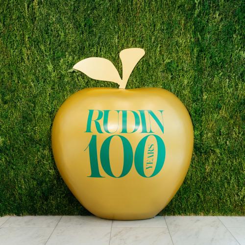 A large golden apple with "Rudin 100 years" written on it sits in front of a plant wall