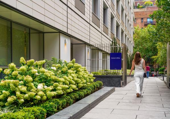 A woman walking along a city sidewalk with a narrow garden and building to her left
