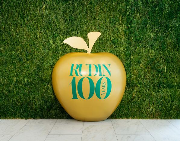 A large golden apple with "Rudin 100 years" written on it sits in front of a plant wall